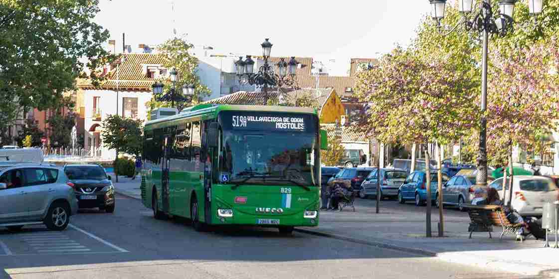 Un autobús verde de la línea 519 circulando por una calle de Villaviciosa de Odón, con árboles y coches aparcados a los lados.