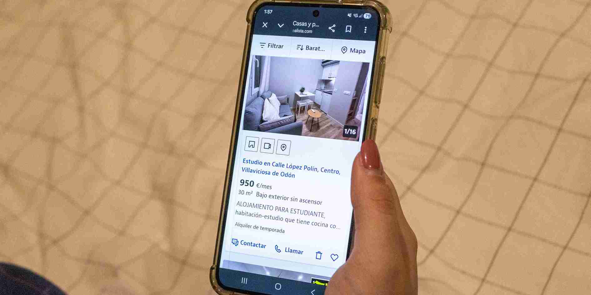 Primer plano de una mano sosteniendo un teléfono móvil que muestra un anuncio de alquiler de un estudio de 30 metros cuadrados por 950 euros al mes en Villaviciosa de Odón.