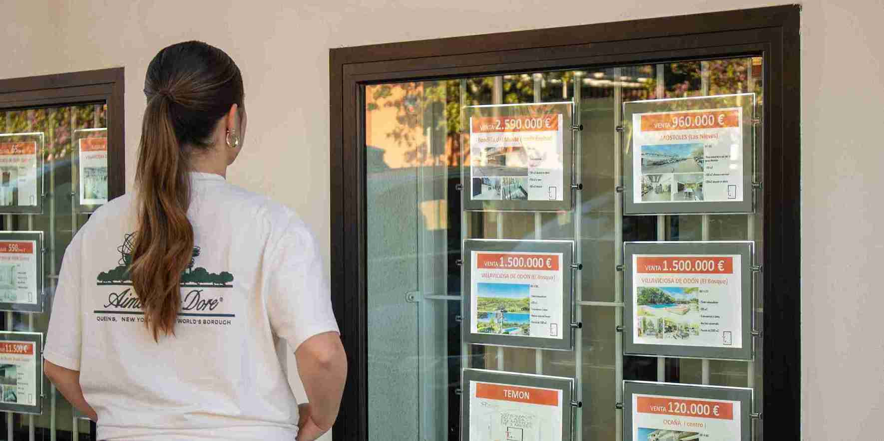 Una mujer joven observa los anuncios de viviendas en el escaparate de una inmobiliaria en Madrid, donde se muestran precios de venta superiores al millón de euros.